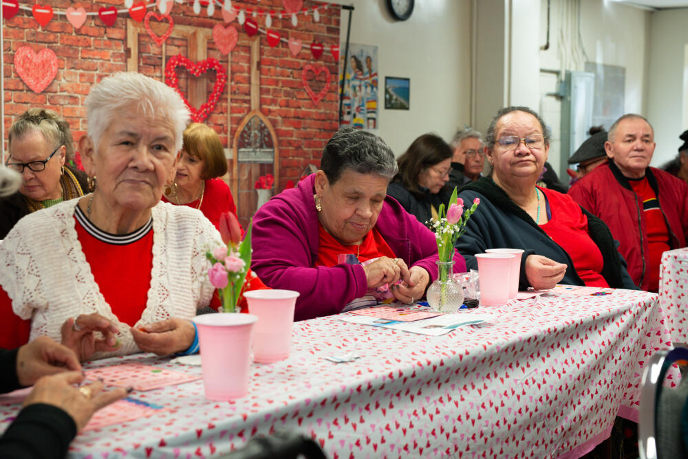 A group of elderly people sit at a long, festively decorated table with pink cups and flower vases, celebrating a Valentine’s-themed event. Red and pink hearts decorate the background wall.