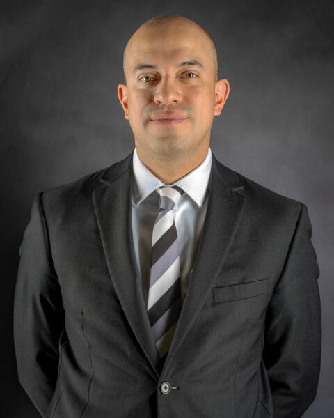 A man in a dark suit, white shirt, and striped tie stands against a plain dark background, looking at the camera with a neutral expression.