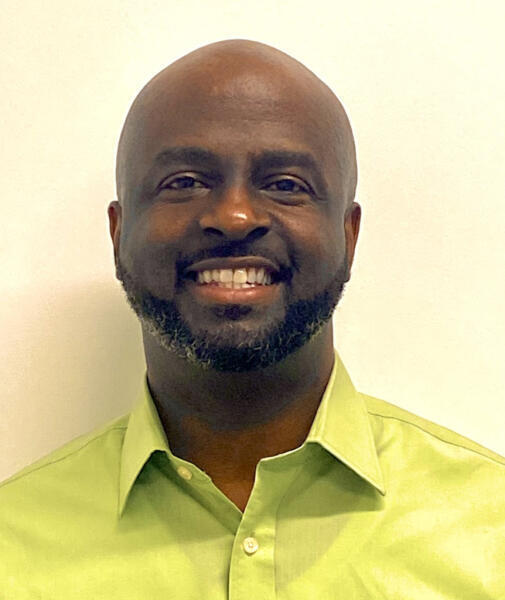 A man with a bald head and trimmed beard, smiling, wearing a light green collared shirt, standing in front of a plain light-colored background.
