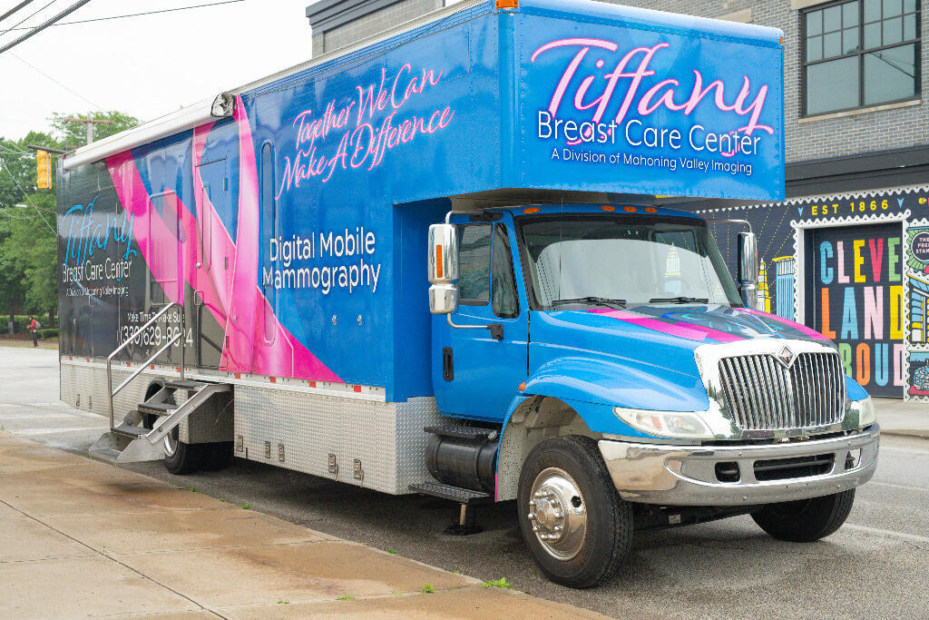 A blue and pink mobile medical truck labeled Tiffany Breast Care Center and Digital Mobile Mammography is parked on a city street near a building with a Cleveland mural.