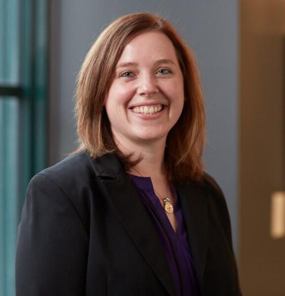 A woman with shoulder-length light brown hair, wearing a black blazer over a dark purple top, smiles while standing indoors near a window with a blurred background.