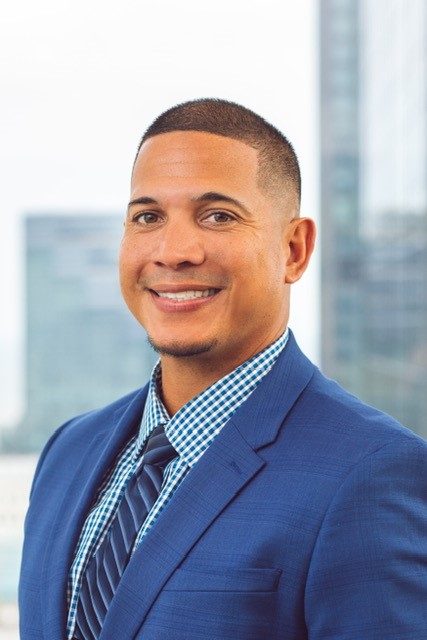 A man in a blue suit, checkered shirt, and striped tie smiles in front of a blurred cityscape background.
