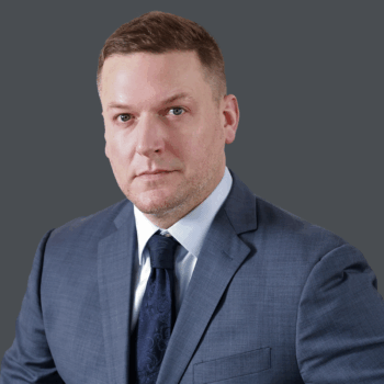 A man in a blue suit and tie poses against a plain gray background, looking directly at the camera with a neutral expression.