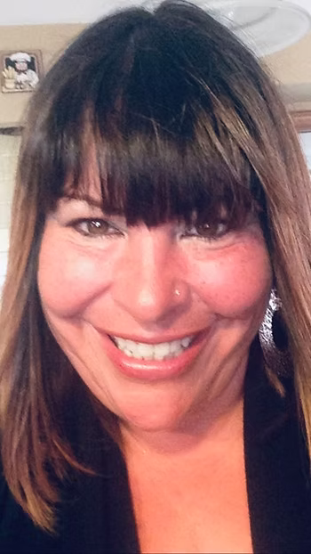 A woman with straight brown hair and bangs smiles at the camera. She is wearing a black top and earrings, and her background includes a beige wall and part of a ceiling light.
